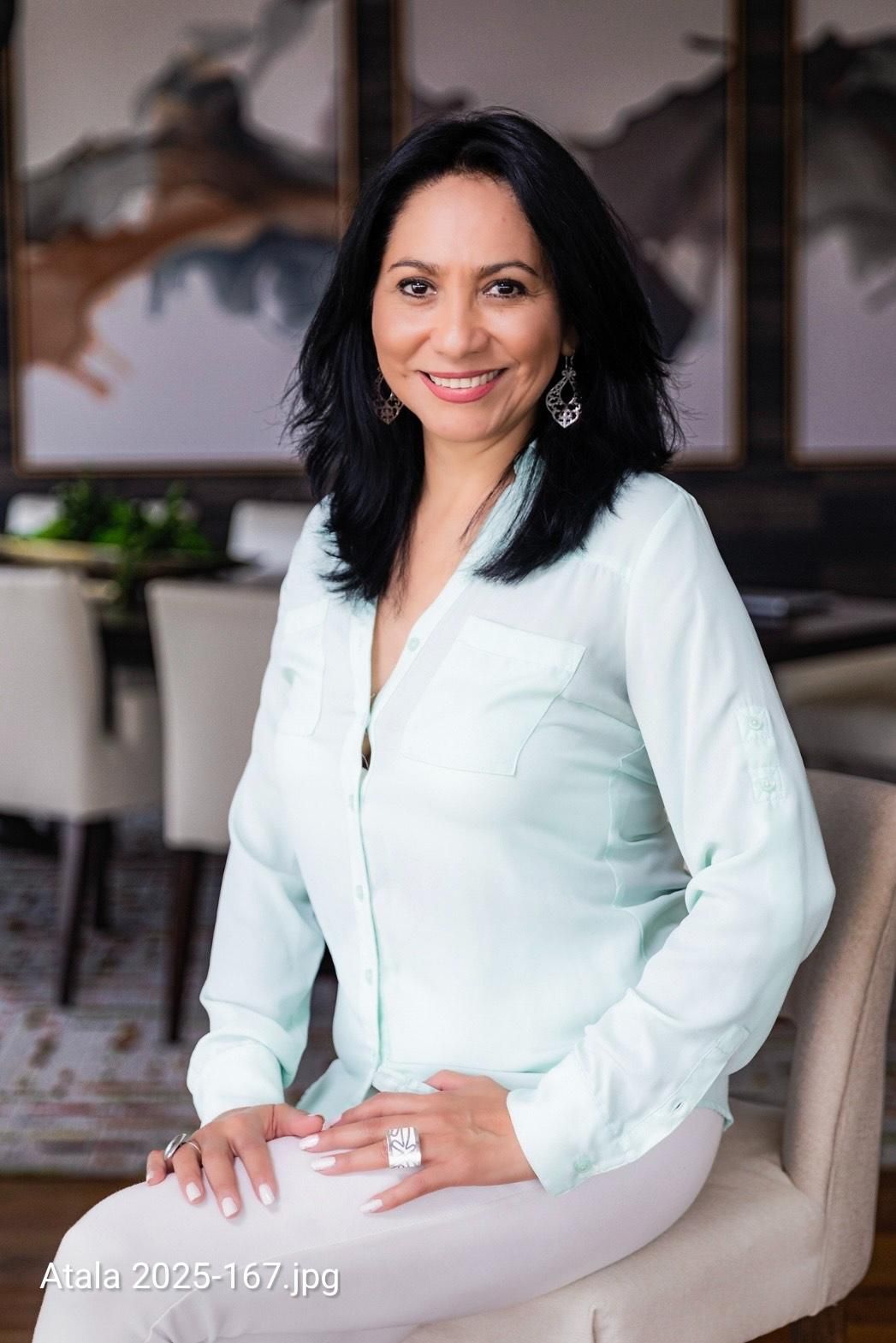 Woman in light blue shirt and white pants smiles, sitting on a chair indoors.