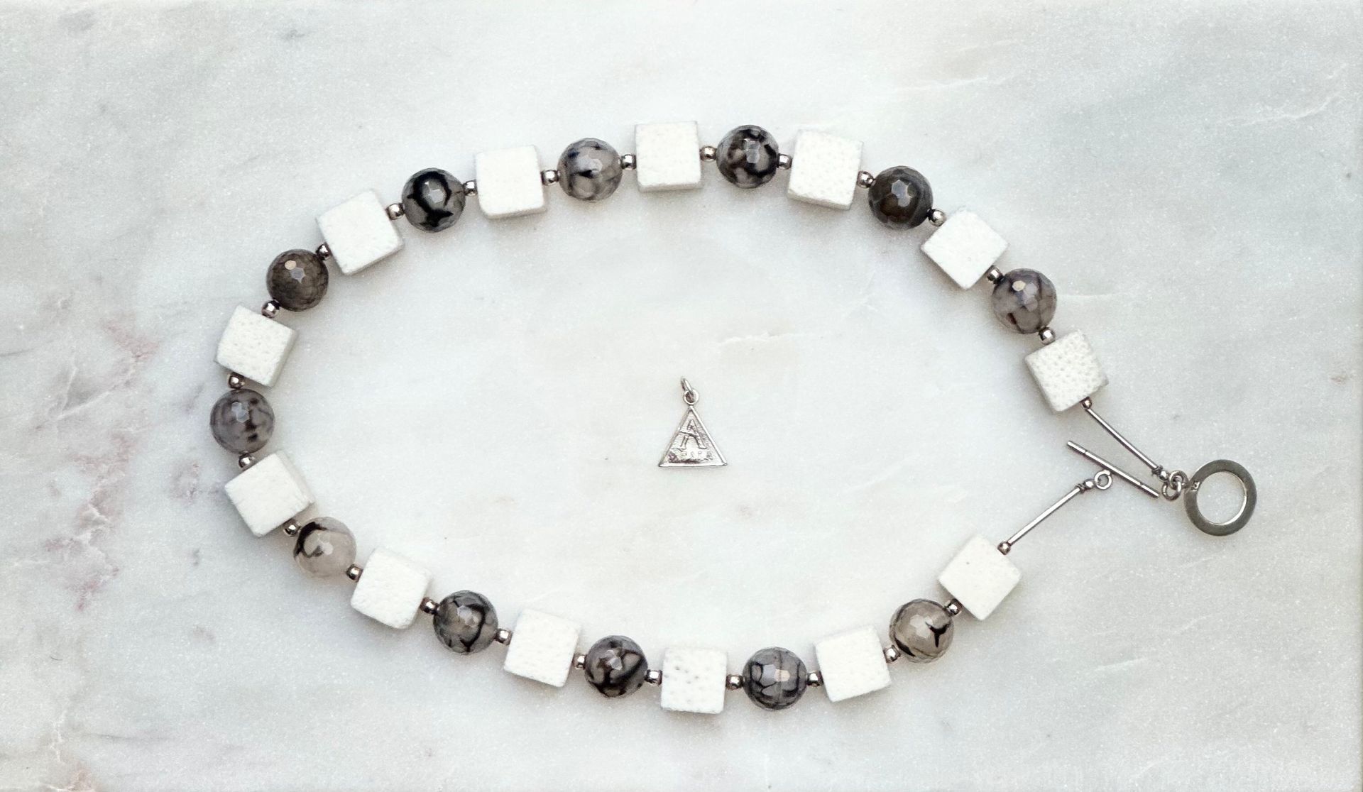 Necklace of white square and grey patterned beads with a triangle pendant on a marbled surface.