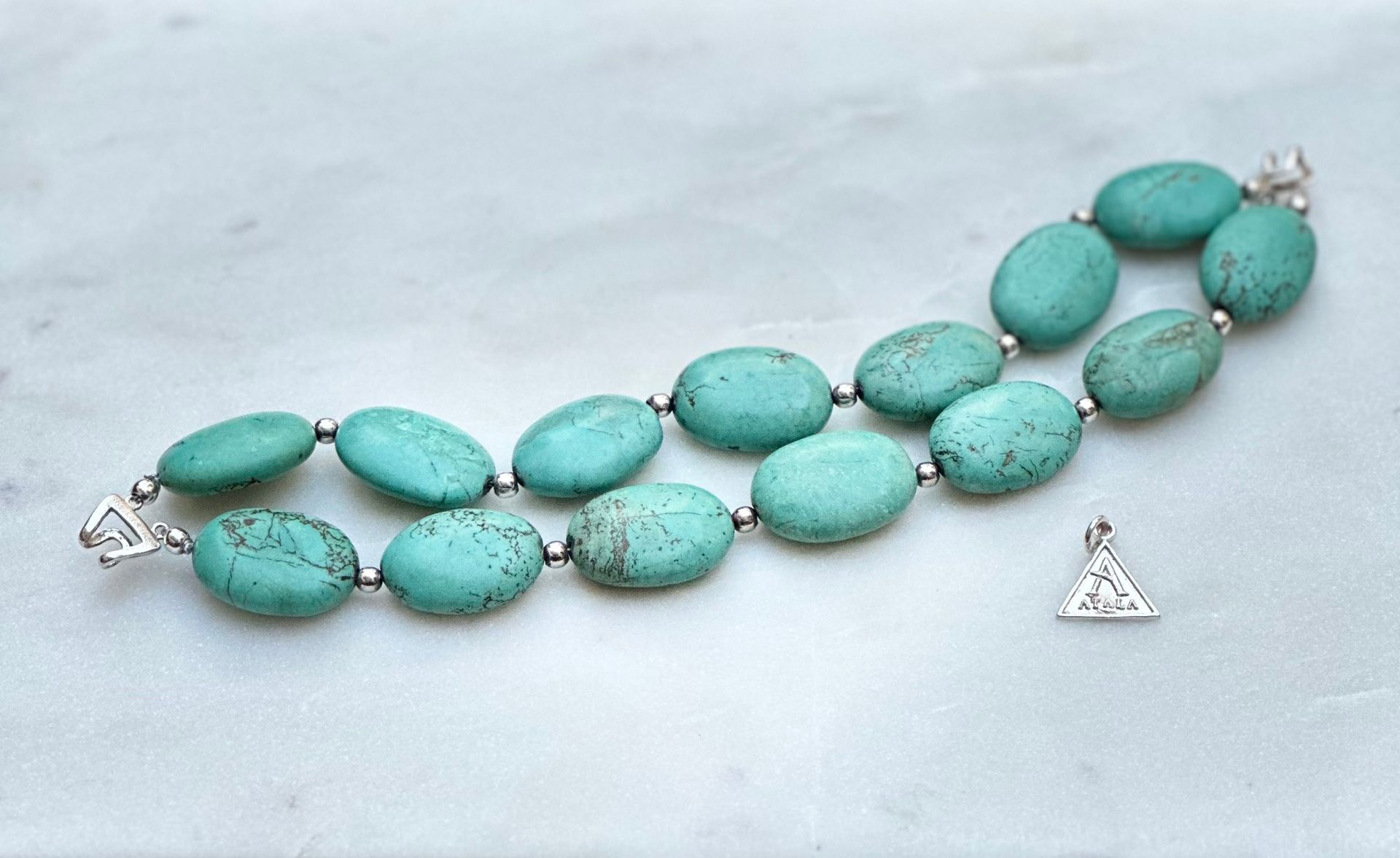 Turquoise stone bracelet with silver accents and small triangular pendant on a marble surface.