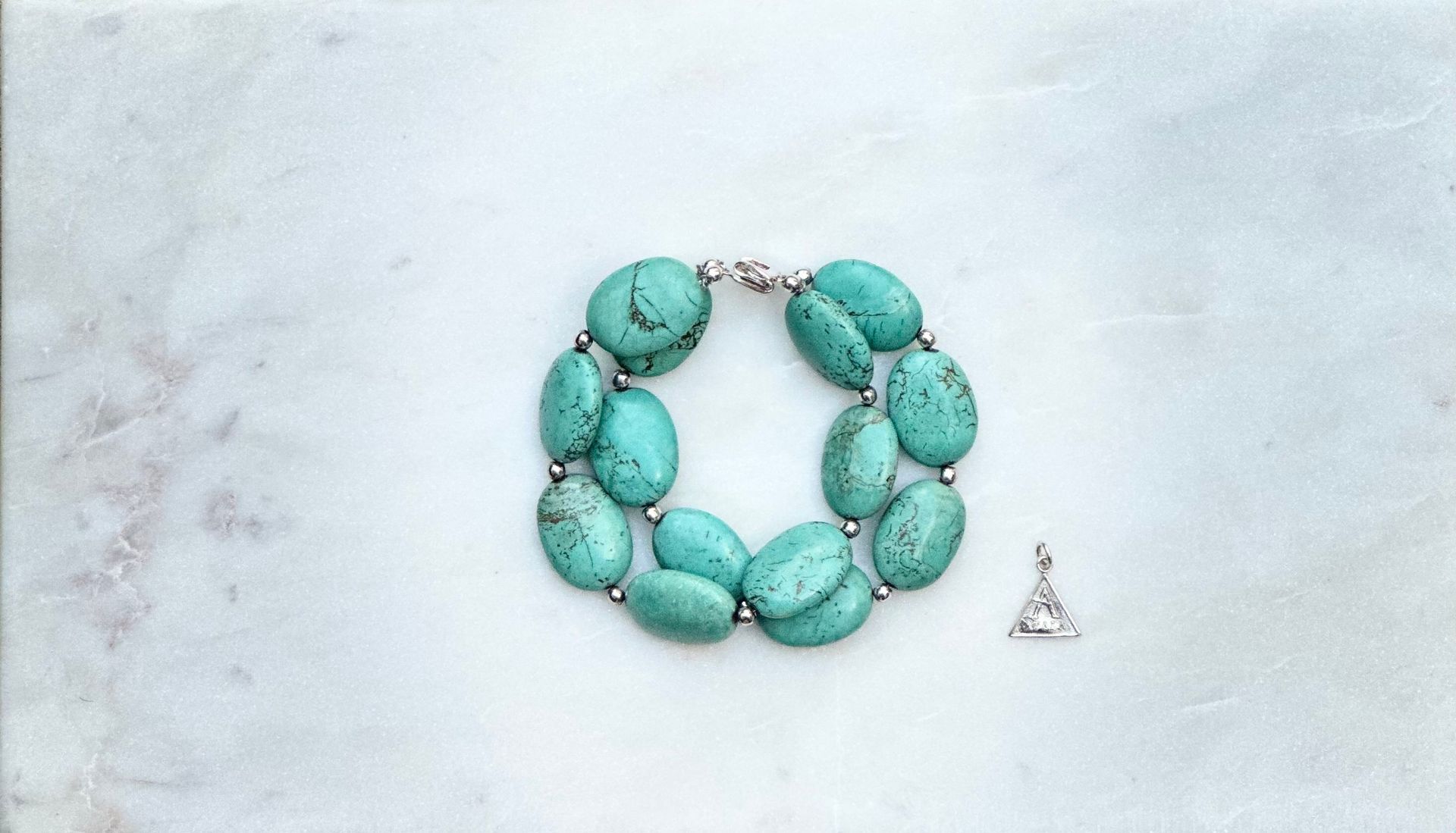Turquoise stone bracelet with silver accents and small triangular pendant on a marble surface.