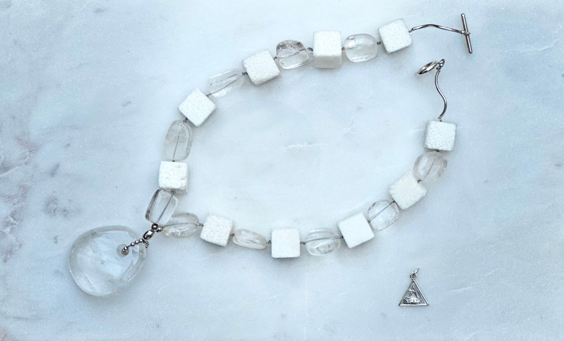 A necklace with a clear teardrop pendant and cube and rounded clear beads. A small triangular charm rests to the side.