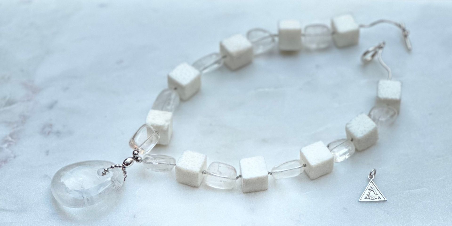 A necklace with a clear teardrop pendant and cube and rounded clear beads. A small triangular charm rests to the side.