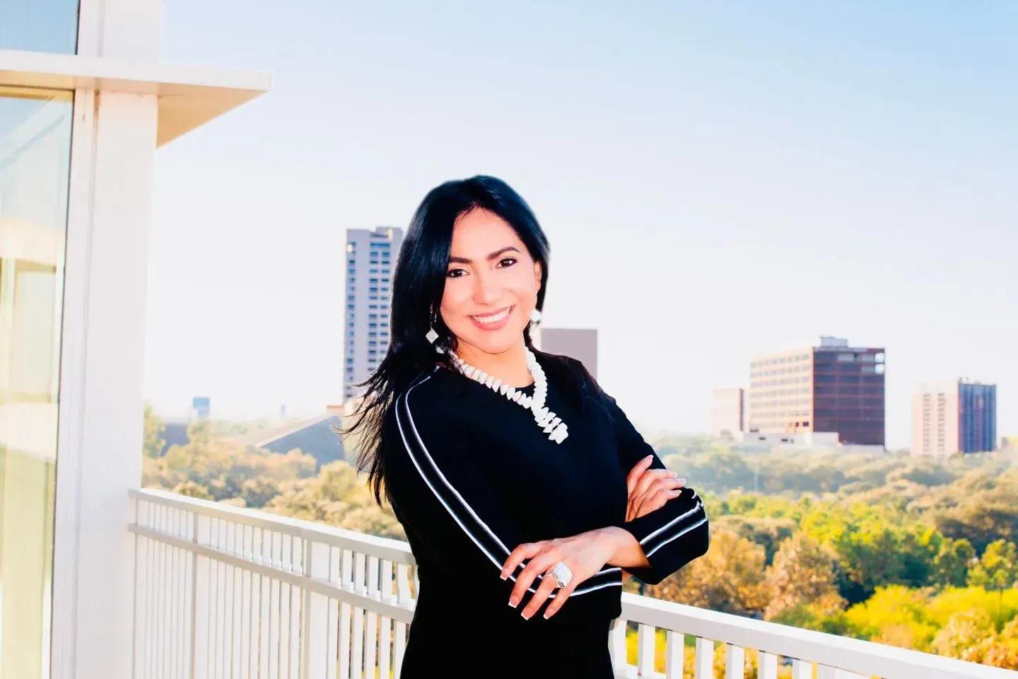 Woman with arms crossed on a balcony, city skyline backdrop. She wears black with jewelry, smiling.