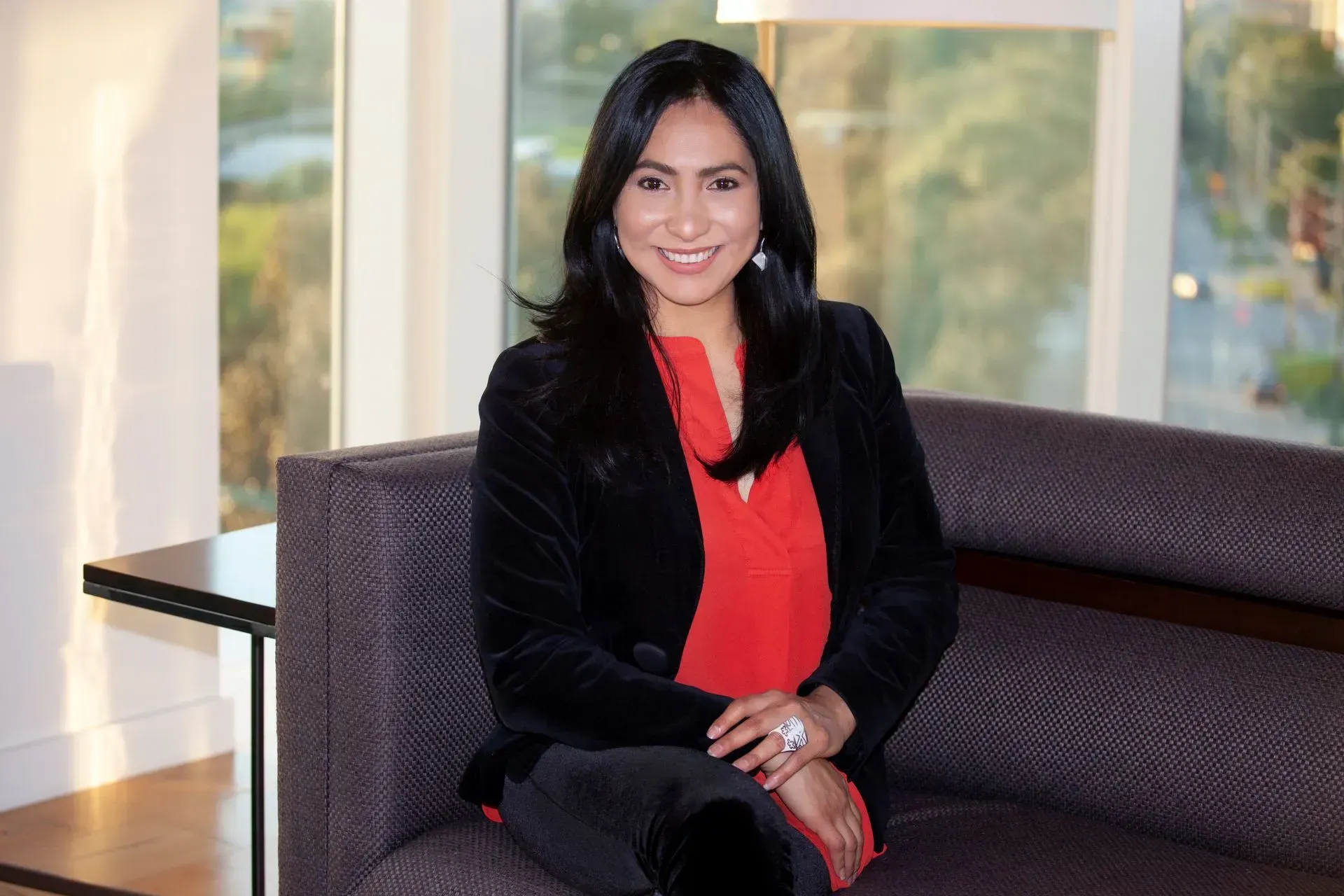 Woman in red blouse and black jacket seated on a gray couch, smiling in front of a window.