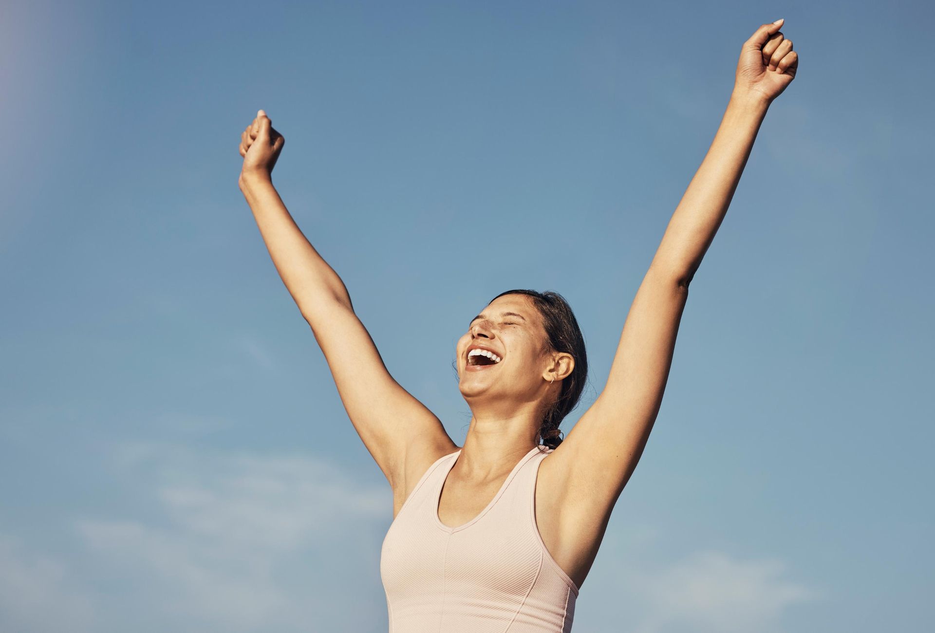 Woman with arms raised, smiling against a blue sky.