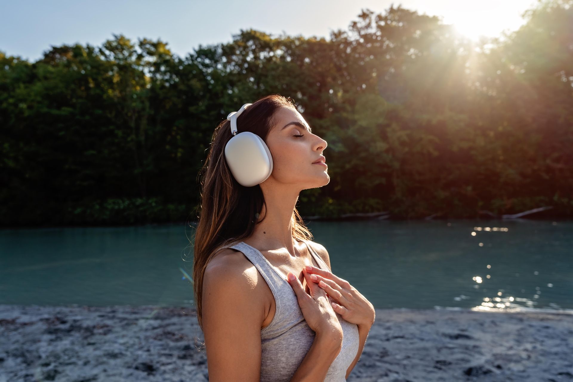 Woman with headphones closes her eyes outdoors, hands on chest, sunlight. River and trees in background.