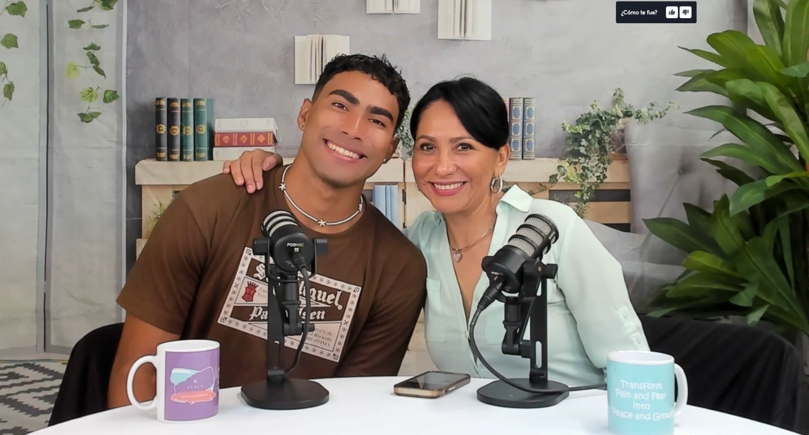 Two people sit at a table with microphones and mugs, smiling at the camera in a studio setting.