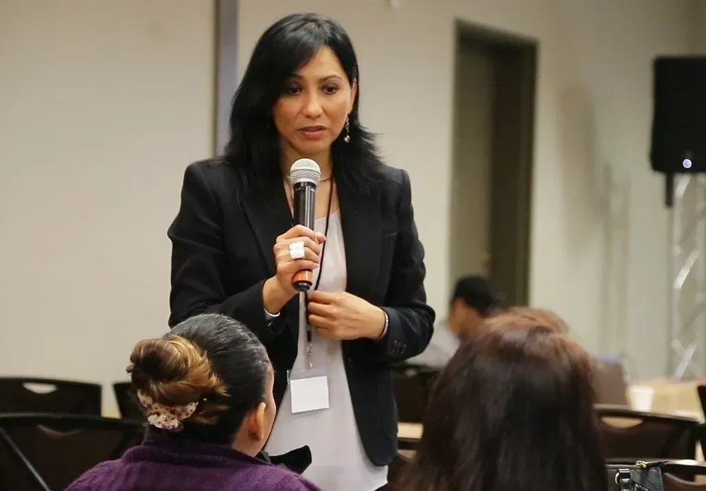 Woman in blazer speaks into microphone at a conference, facing an audience.