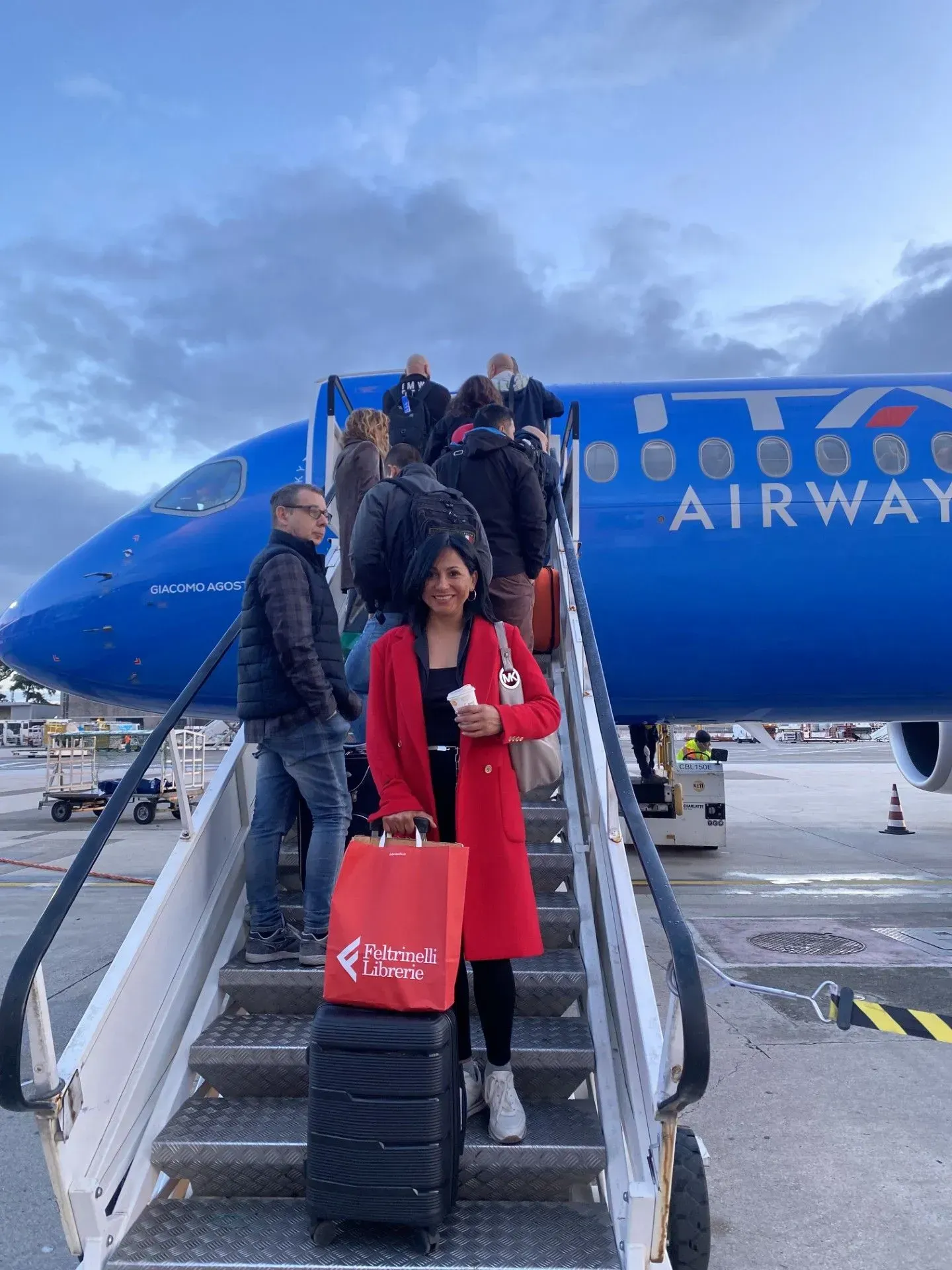 People boarding a blue ITA Airways plane via stairs. Woman in red coat smiles, holding luggage and coffee. Cloudy sky.