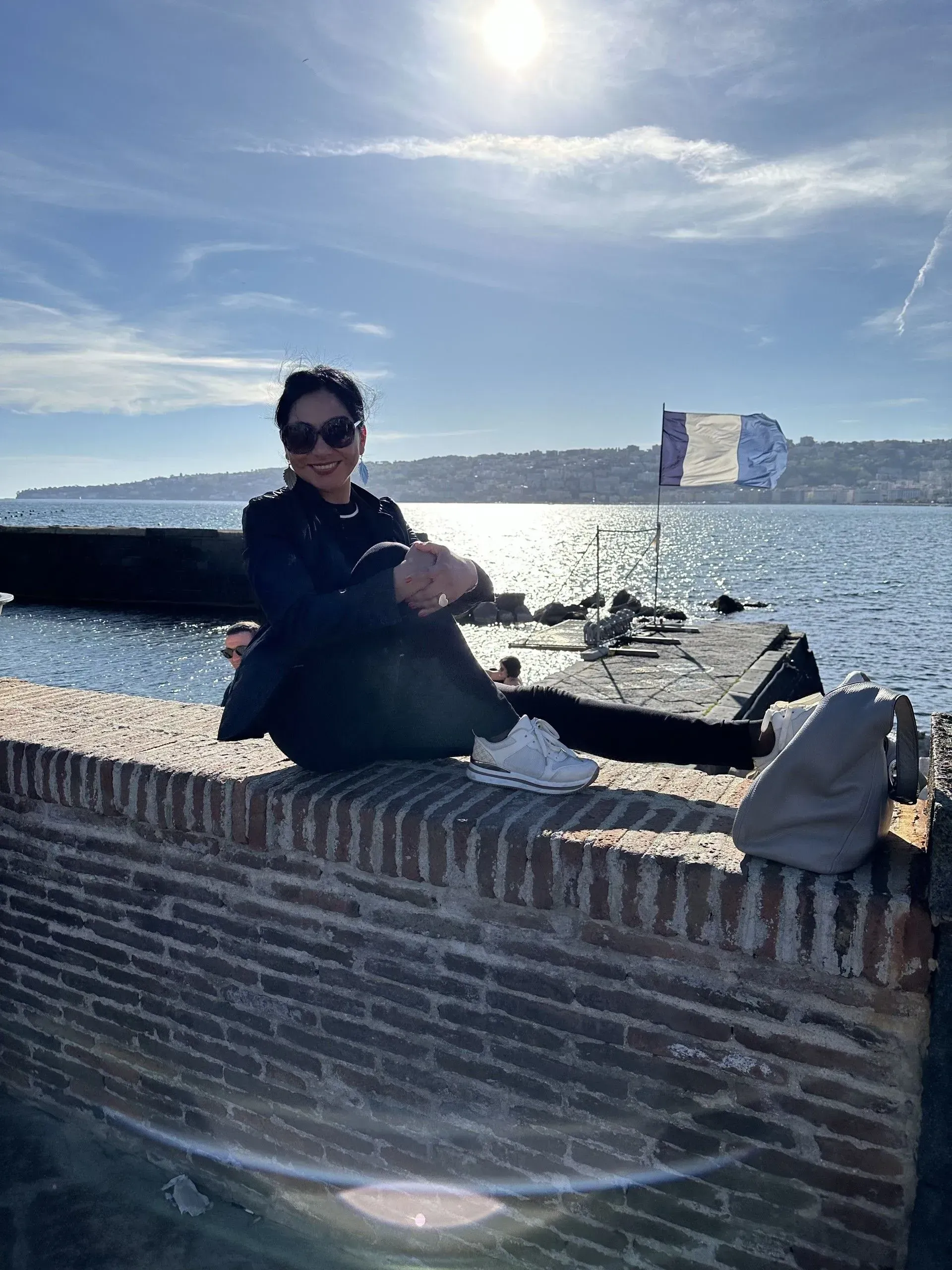 Person sitting on a brick wall by the sea, bright sunlight.