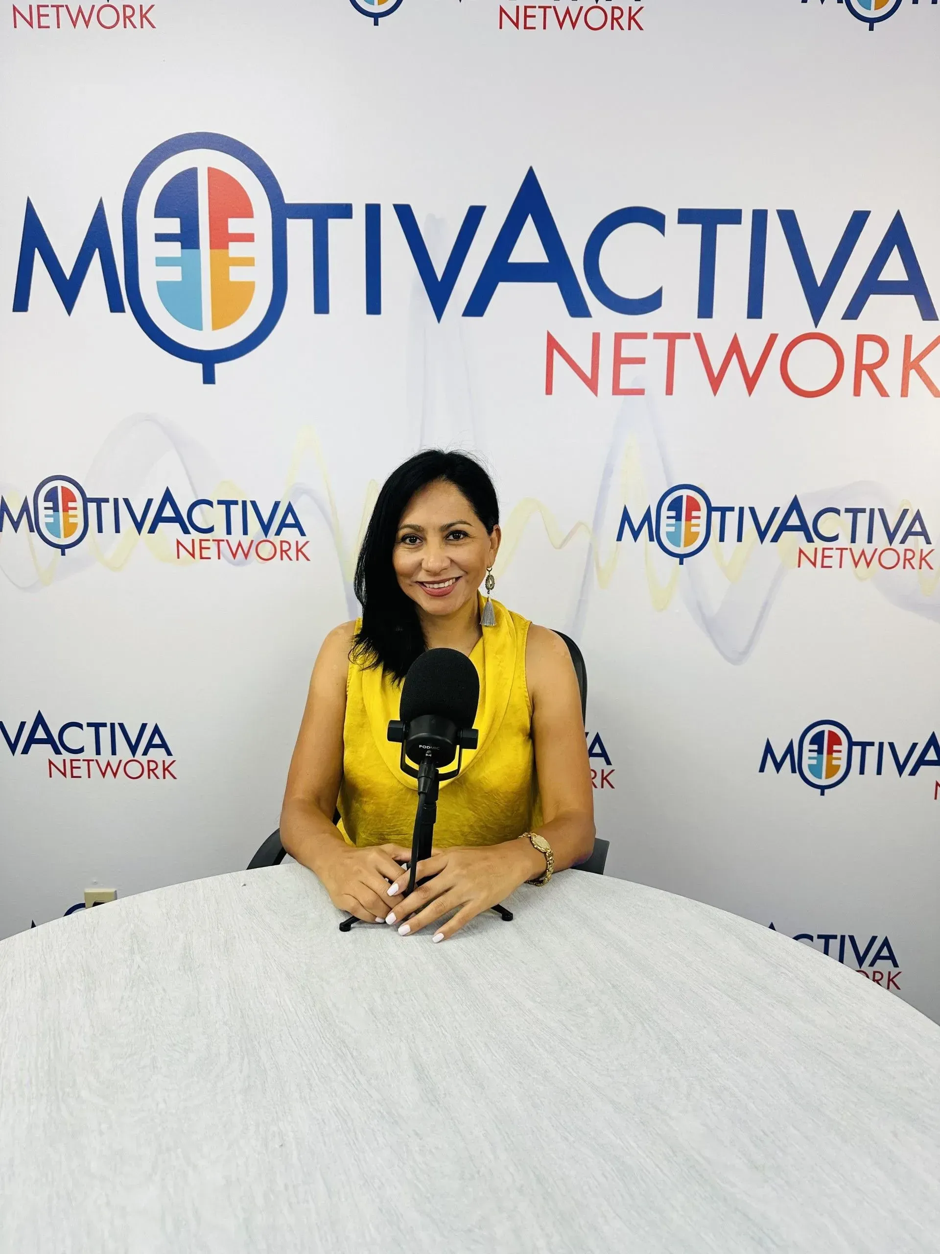 Woman in yellow top sits at a table with microphone, logo backdrop: MotivActiva Network.