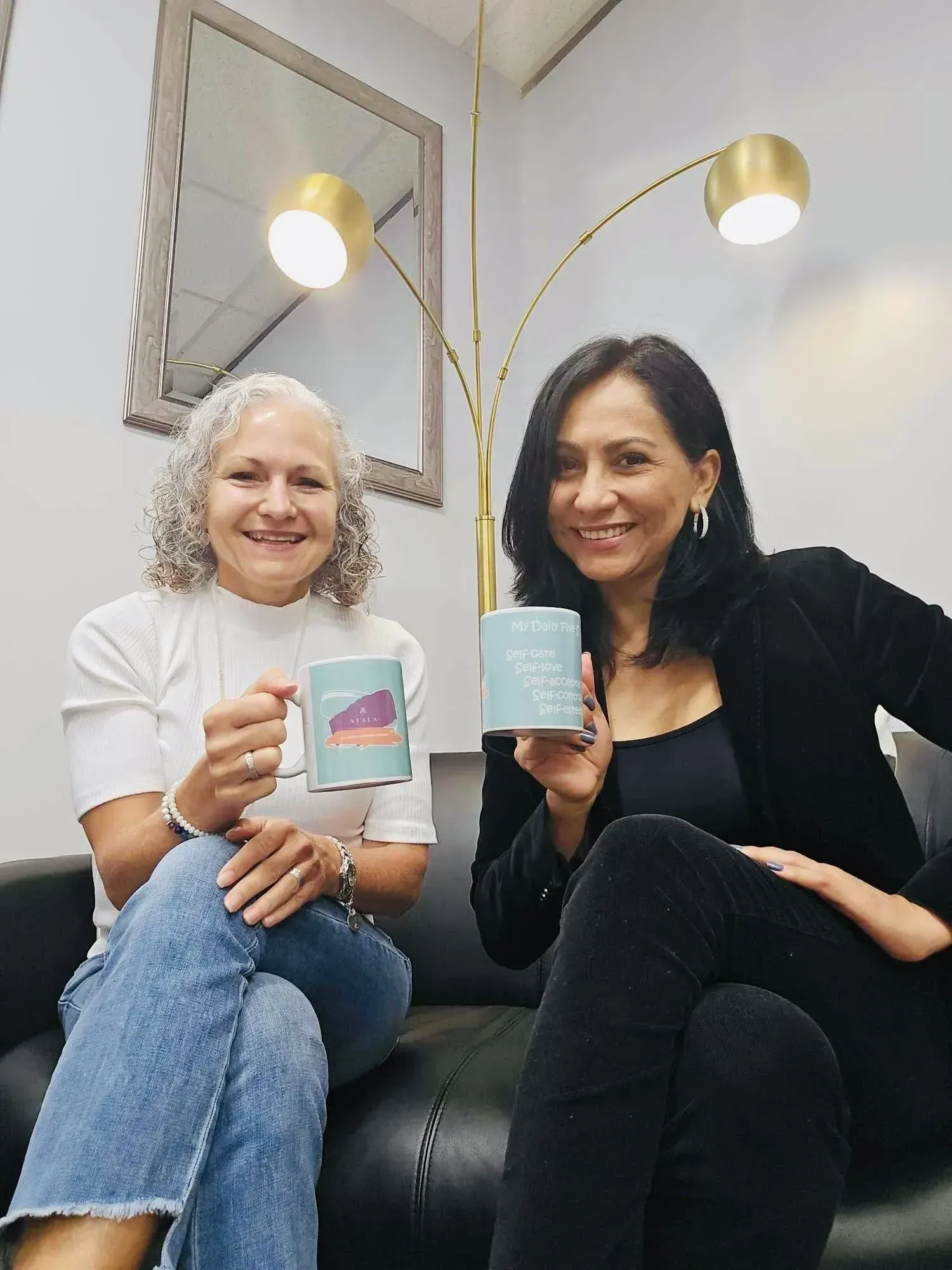 Two women seated, holding mugs. One in jeans, one in black; smiling. Modern office with gold lamp.