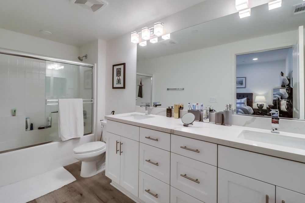 White bathroom with double vanity, bathtub, large mirror, and open doorway to a bedroom.