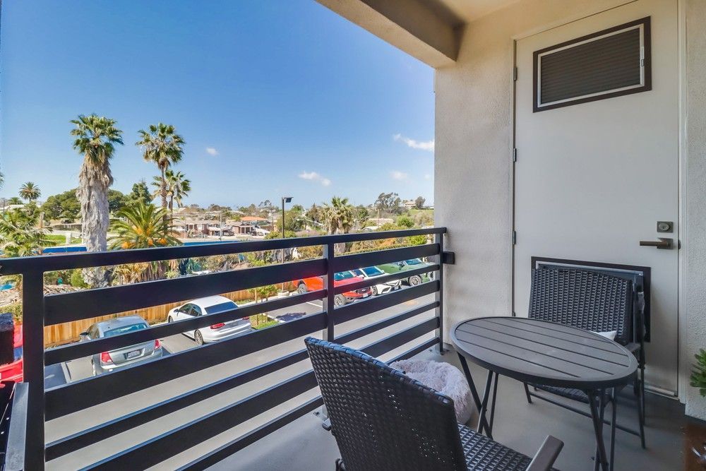 Balcony view with table, chairs, railing, and palm trees. Sunny day with parked cars visible.