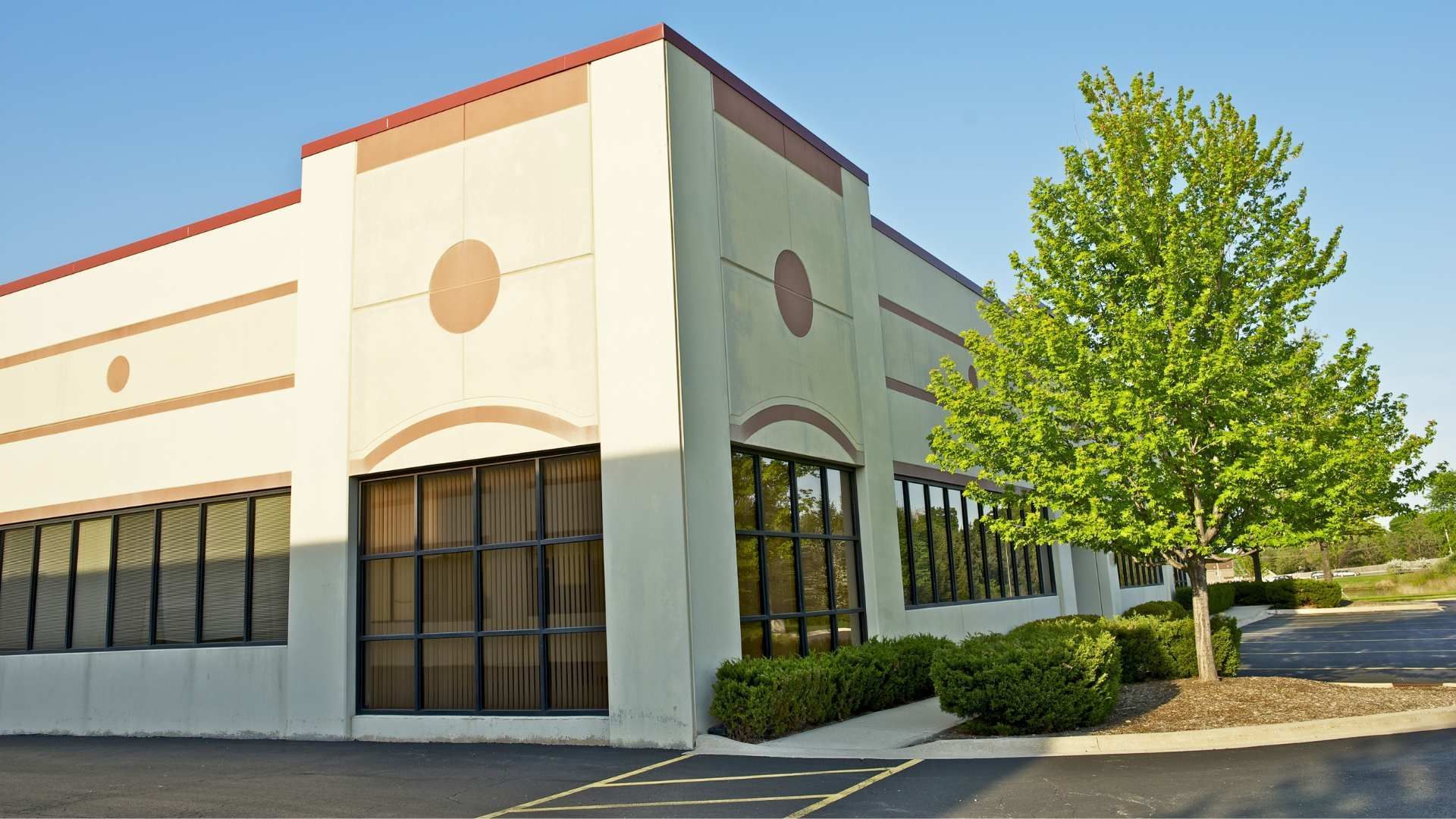 Exterior of a beige commercial building with dark windows and a tree-lined parking lot under clear skies