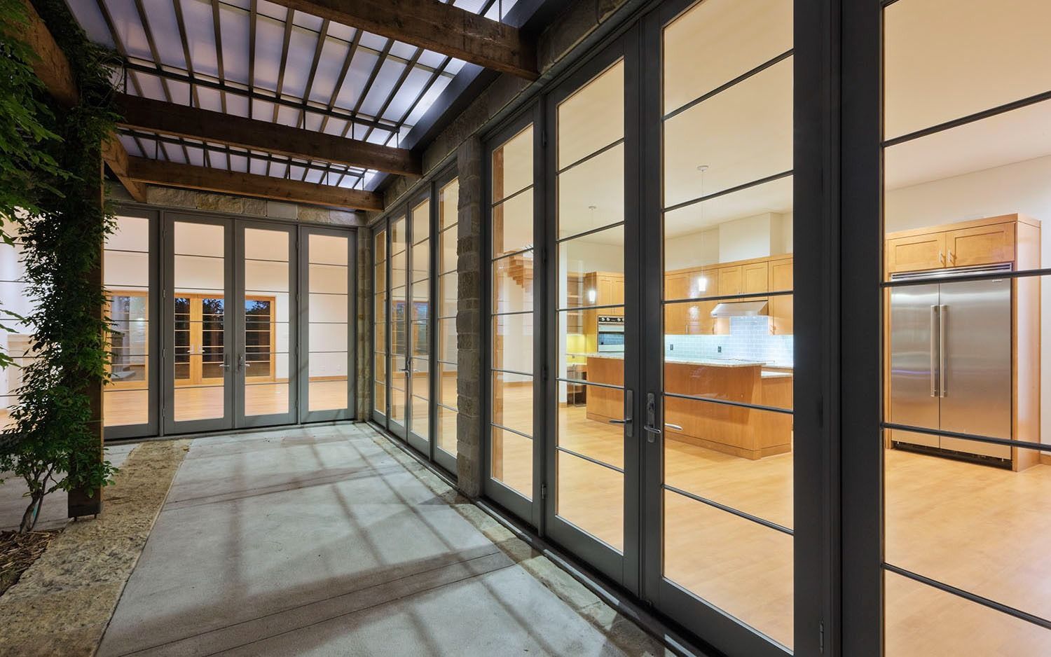 Glass corridor beside a modern indoor kitchen, with black-framed windows and warm wood flooring