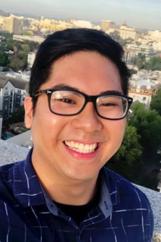 Smiling person wearing glasses and a blue shirt, taking a selfie with city buildings in the background