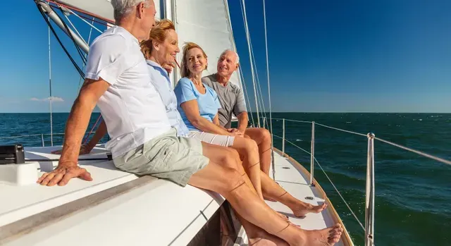 Three people sit on a sailboat deck, relaxing in bright sun on calm blue water.