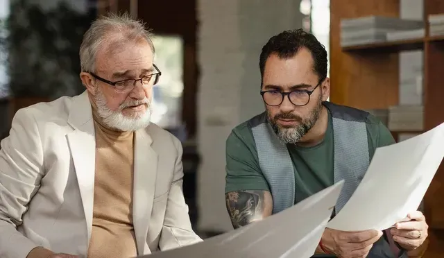 Two coworkers review documents together at a desk in a bright office, one in a white jacket and one in a green sweater.