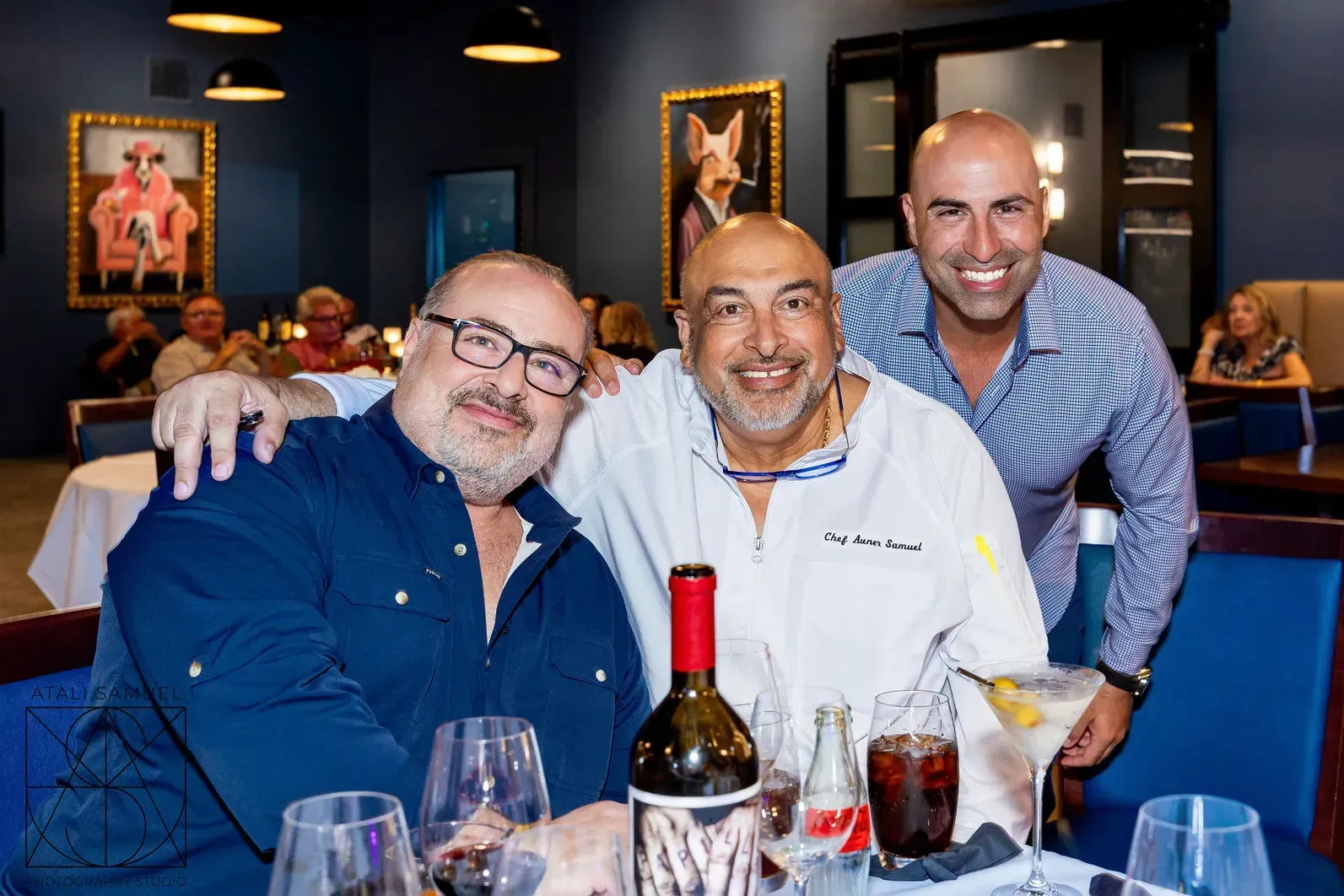 Three men at a restaurant posing together. One in chef's whites, bottle of wine on the table, paintings on the wall.