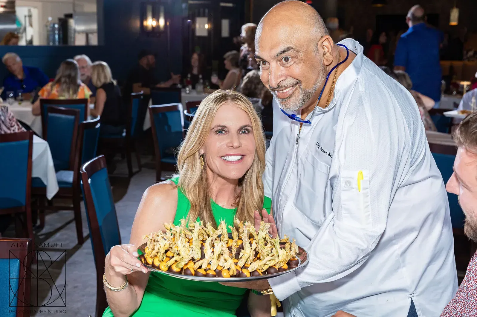 Woman in green dress smiles, holding a tray of loaded fries, chef beside her, restaurant setting.