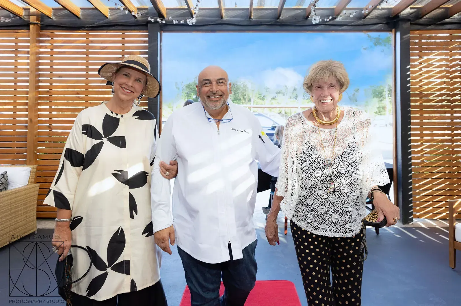 Three people smiling, walking together, outdoors. Middle person has arm around each person.