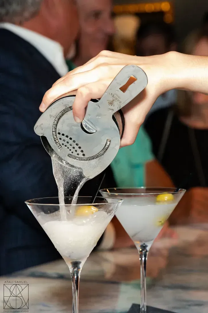 Bartender pouring cocktails with a strainer into two martini glasses, garnished with olives.