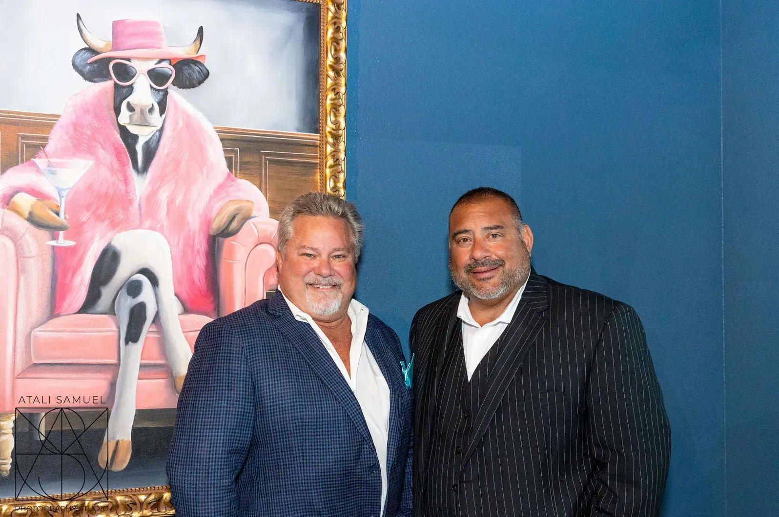 Two men pose next to a painting of a cow in a pink coat and sunglasses, holding a martini.