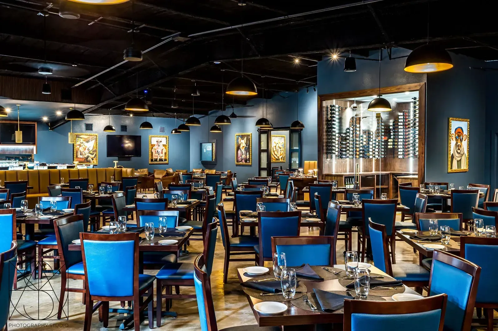 Restaurant interior with blue chairs, dark tables, and artwork on dark blue walls; wine display.
