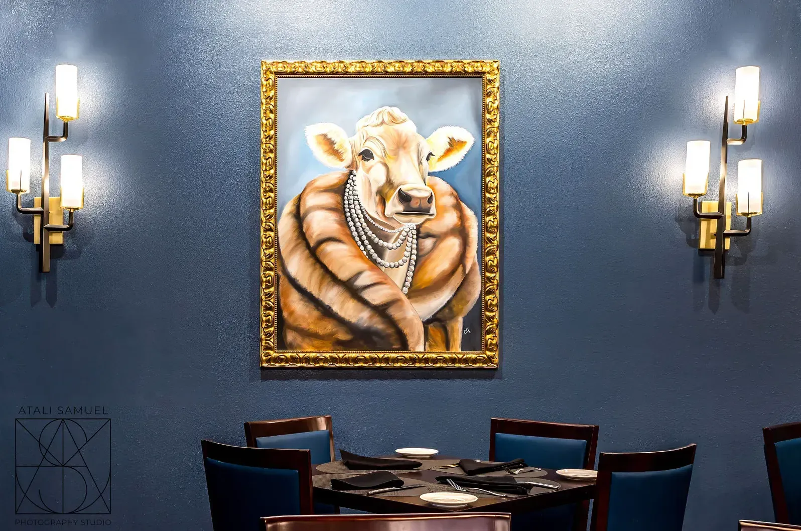 Restaurant dining area with a painting of a cow wearing a necklace. Dark blue walls, sconce lighting, and a table set for dining.