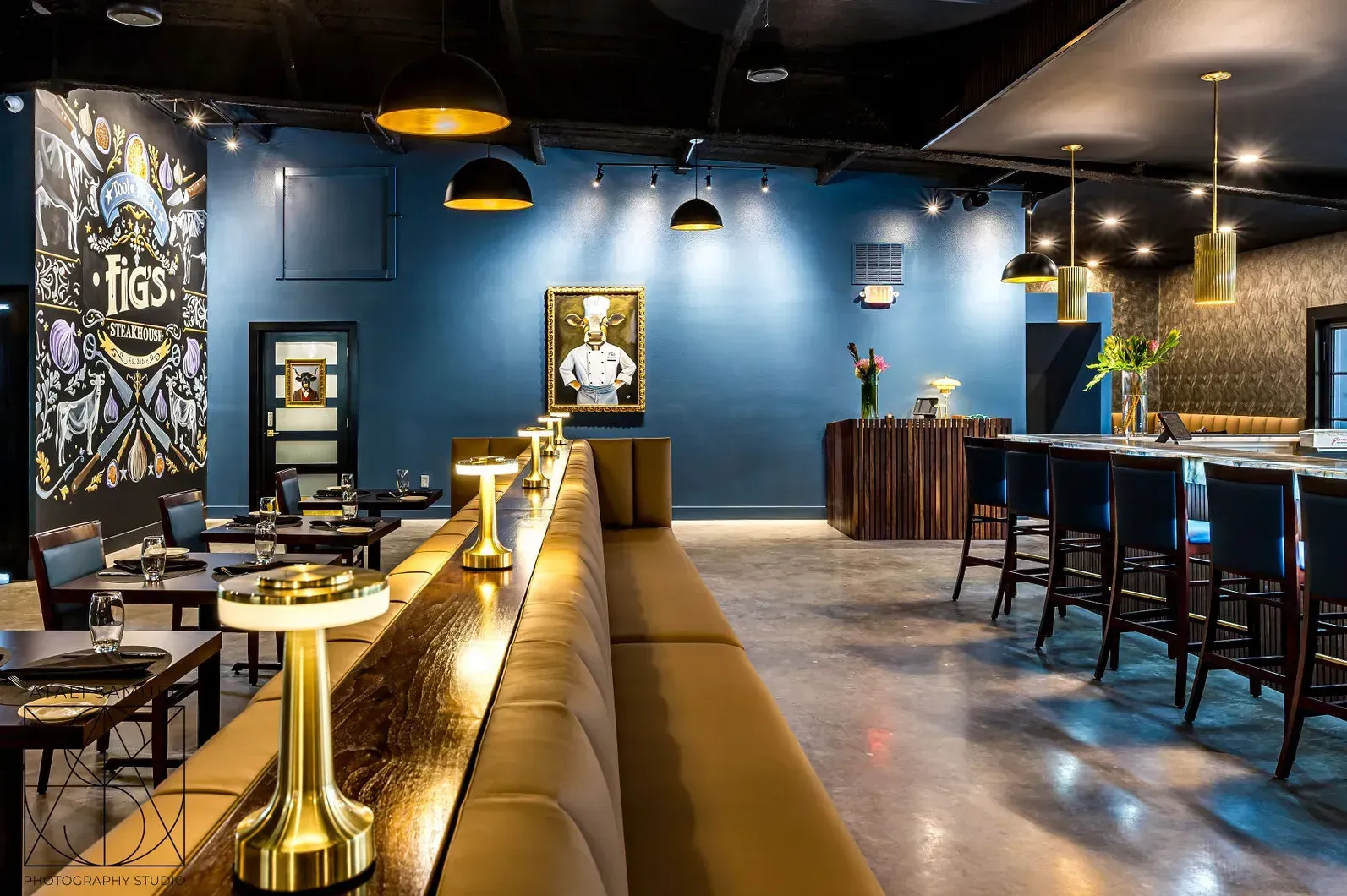 Interior of a modern restaurant: blue walls, gold accents, bar seating, booth with gold lamps, artwork, and a bar.