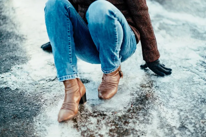 Person squatting, wearing jeans and brown boots, losing balance on icy ground. Black gloves on one hand.