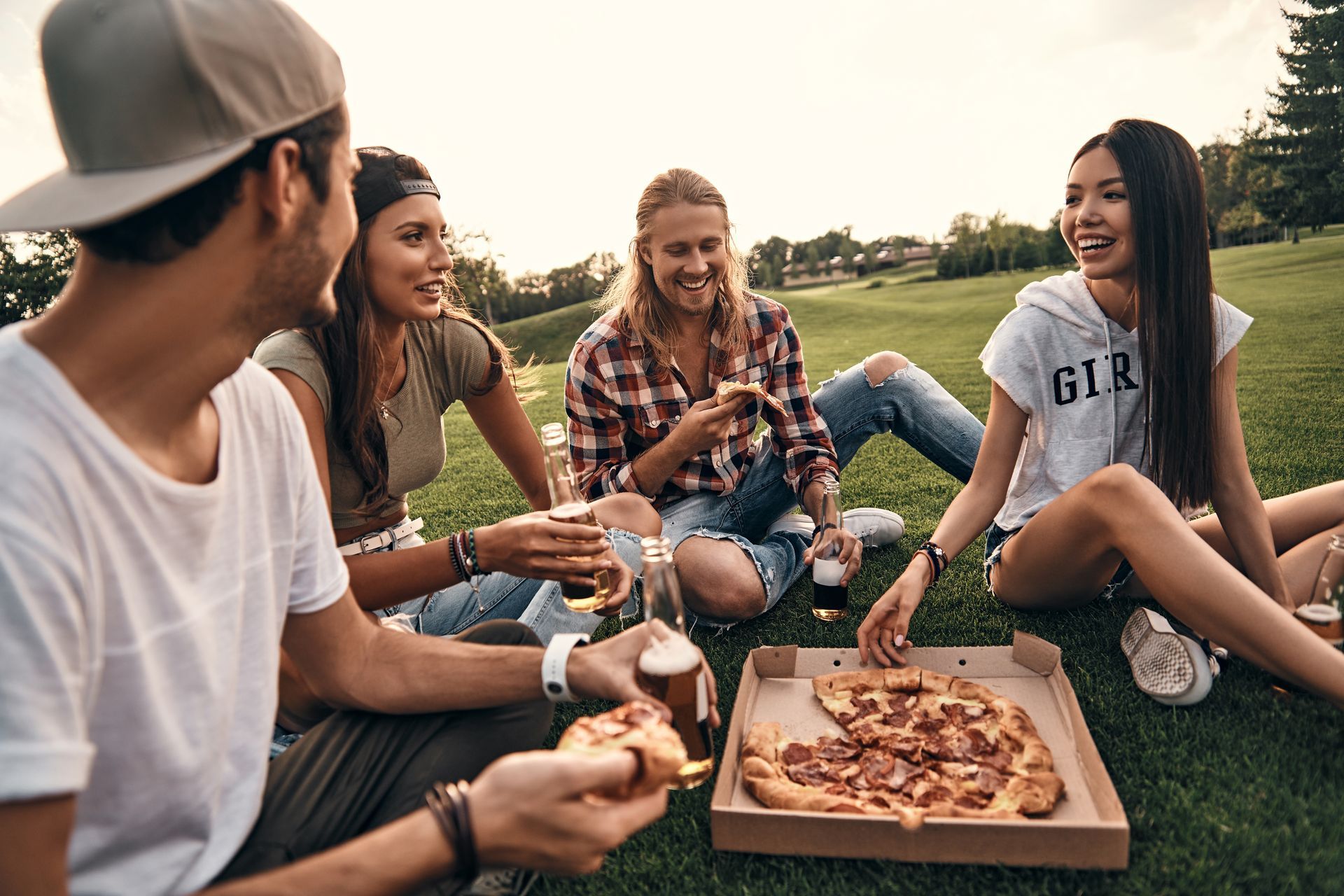 A group of young people are sitting on the grass eating pizza and drinking beer.
