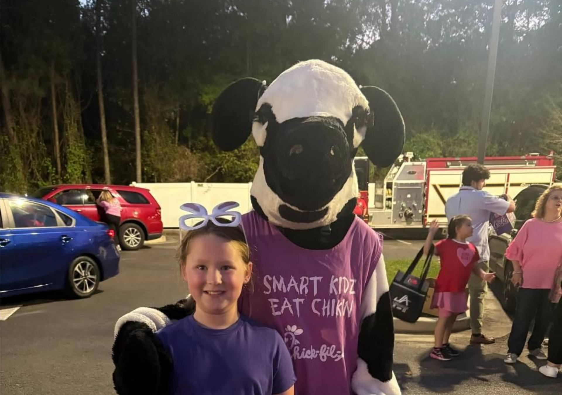 Person posing with a black-and-white cow mascot in purple shirts at an outdoor event, near parked cars.