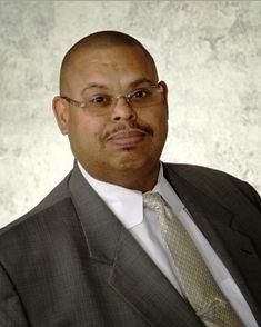 A professional headshot of a person wearing glasses, a grey suit, a white collared shirt, and a light-patterned tie.