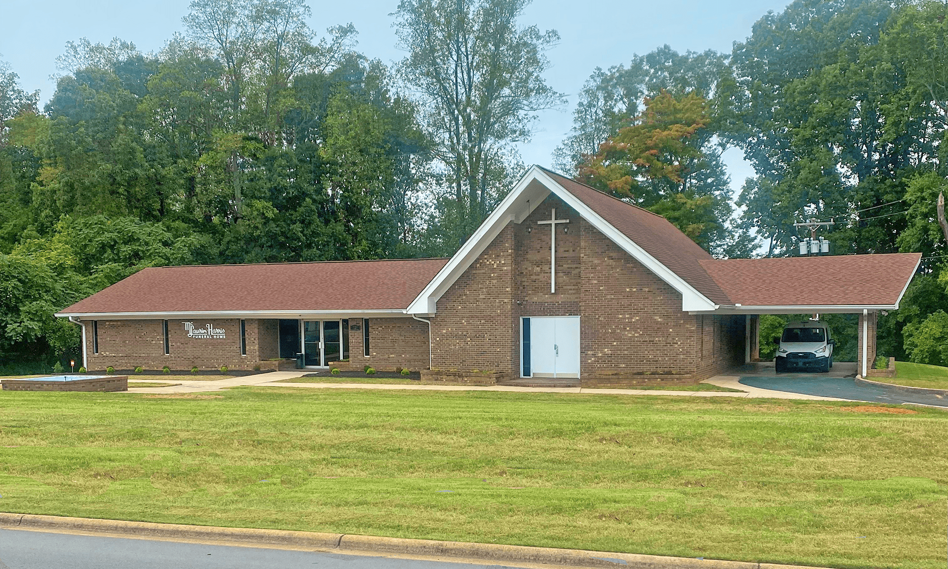 A one-story brick church building with a steep gabled entrance featuring a cross, situated on a grassy lawn with trees.