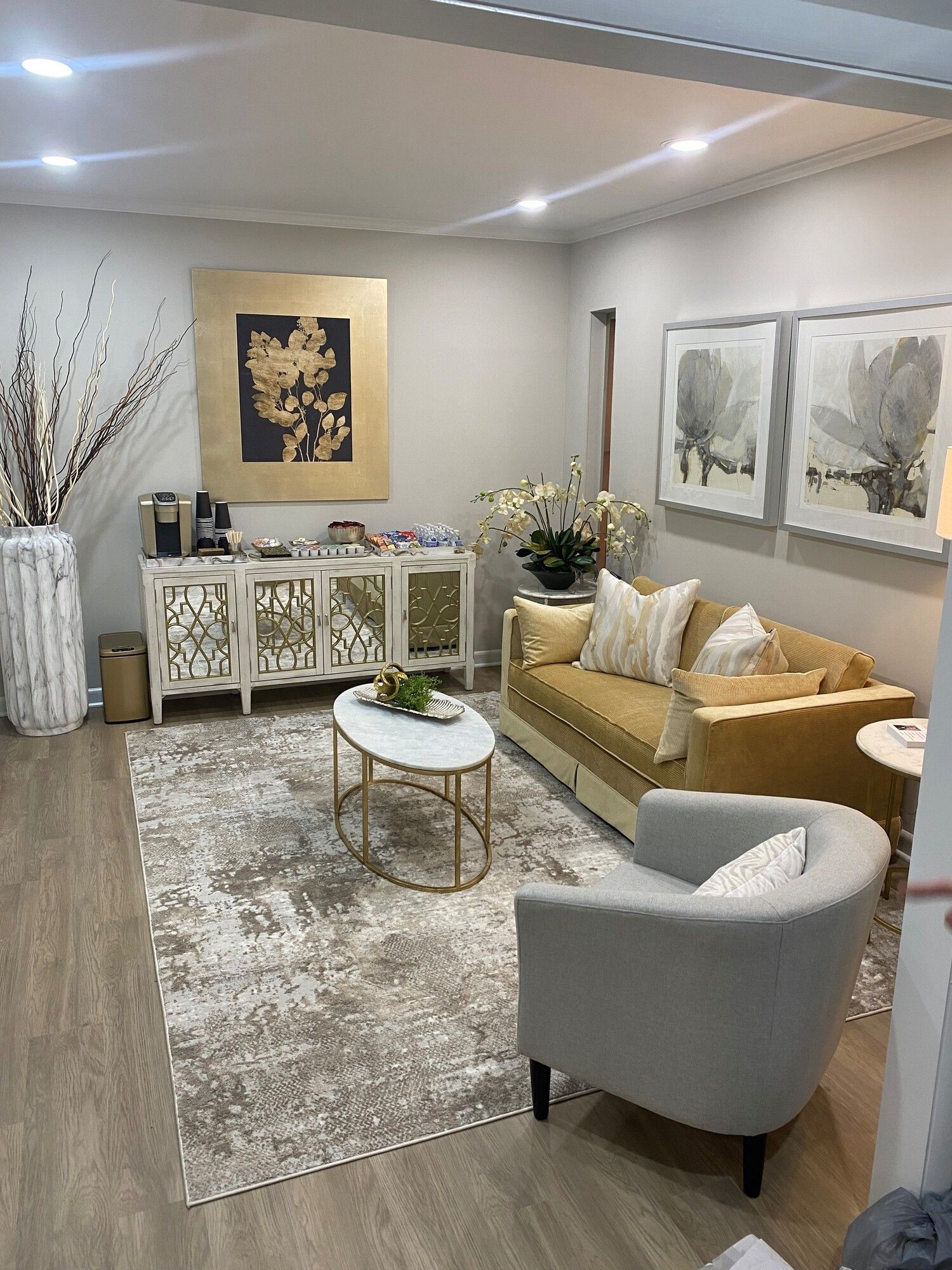 A modern seating area with a gold sofa, gray armchair, white oval coffee table, patterned rug, and a decorative sideboard.