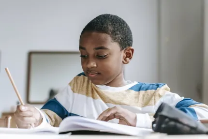 Boy focused on writing in notebook with a pencil at a desk. Wearing striped shirt.