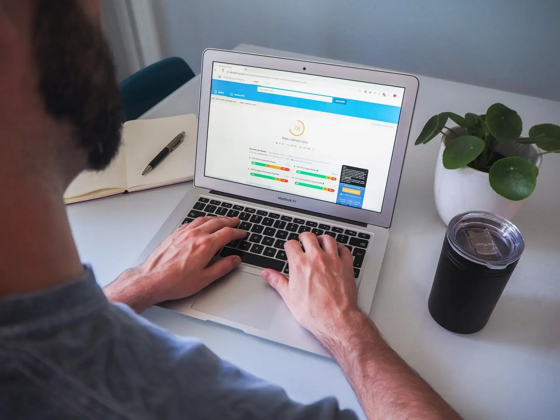 Person typing on a laptop at a desk with a notebook, plant, and drink nearby.