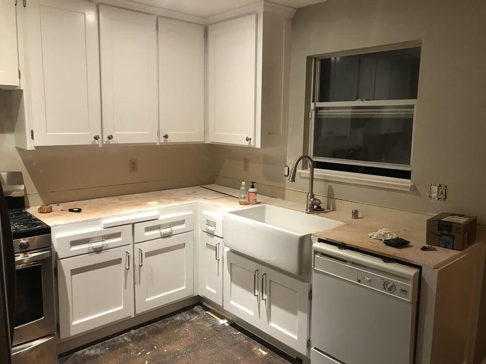 Small beige kitchen with white cabinets, stove, sink, dishwasher, and a window over the counter.