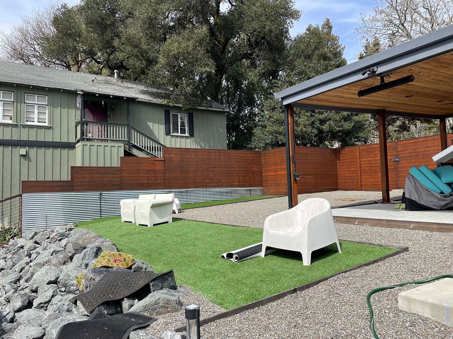 Backyard patio with artificial turf, white chairs, rocks, and a covered seating area beside a house