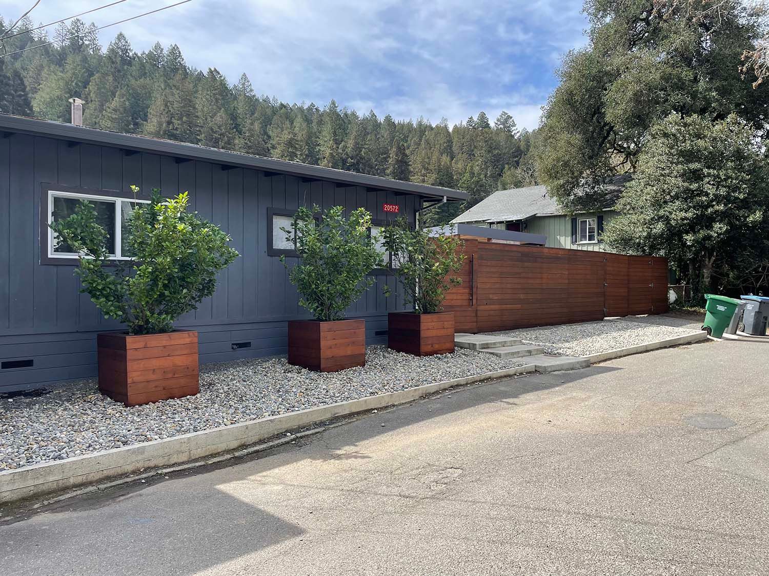 Single-story dark gray house with potted plants, red fence, and gravel driveway under a partly cloudy sky
