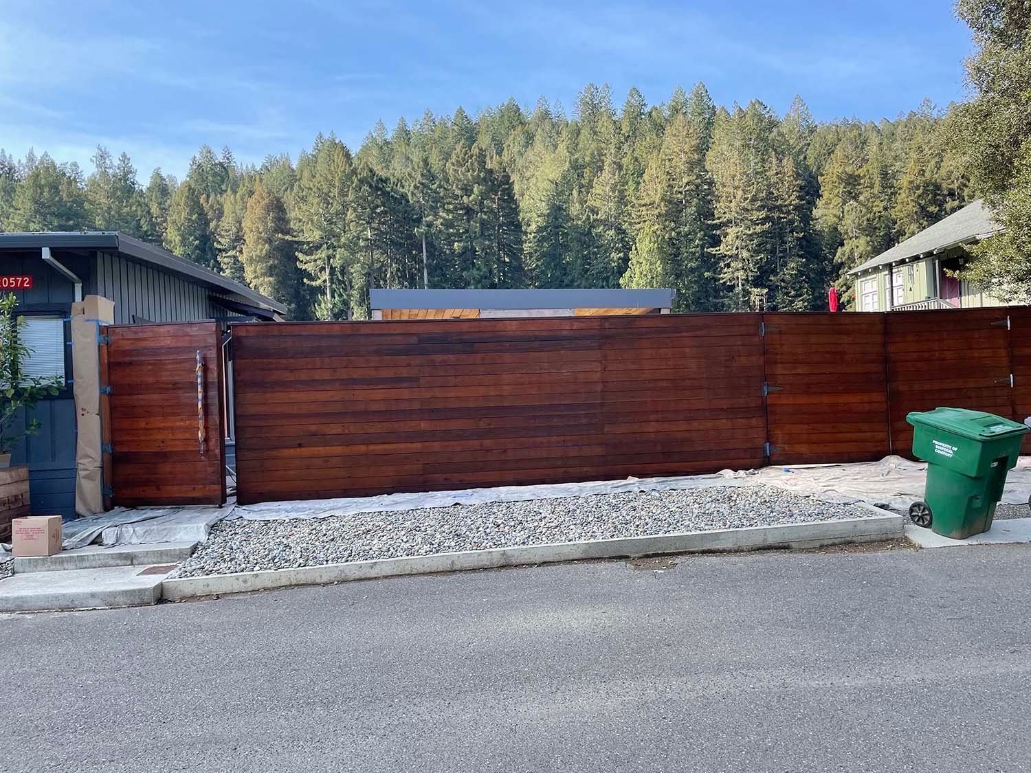 Wooden privacy fence across a gravel front yard, with a green trash bin beside a road and trees behind.