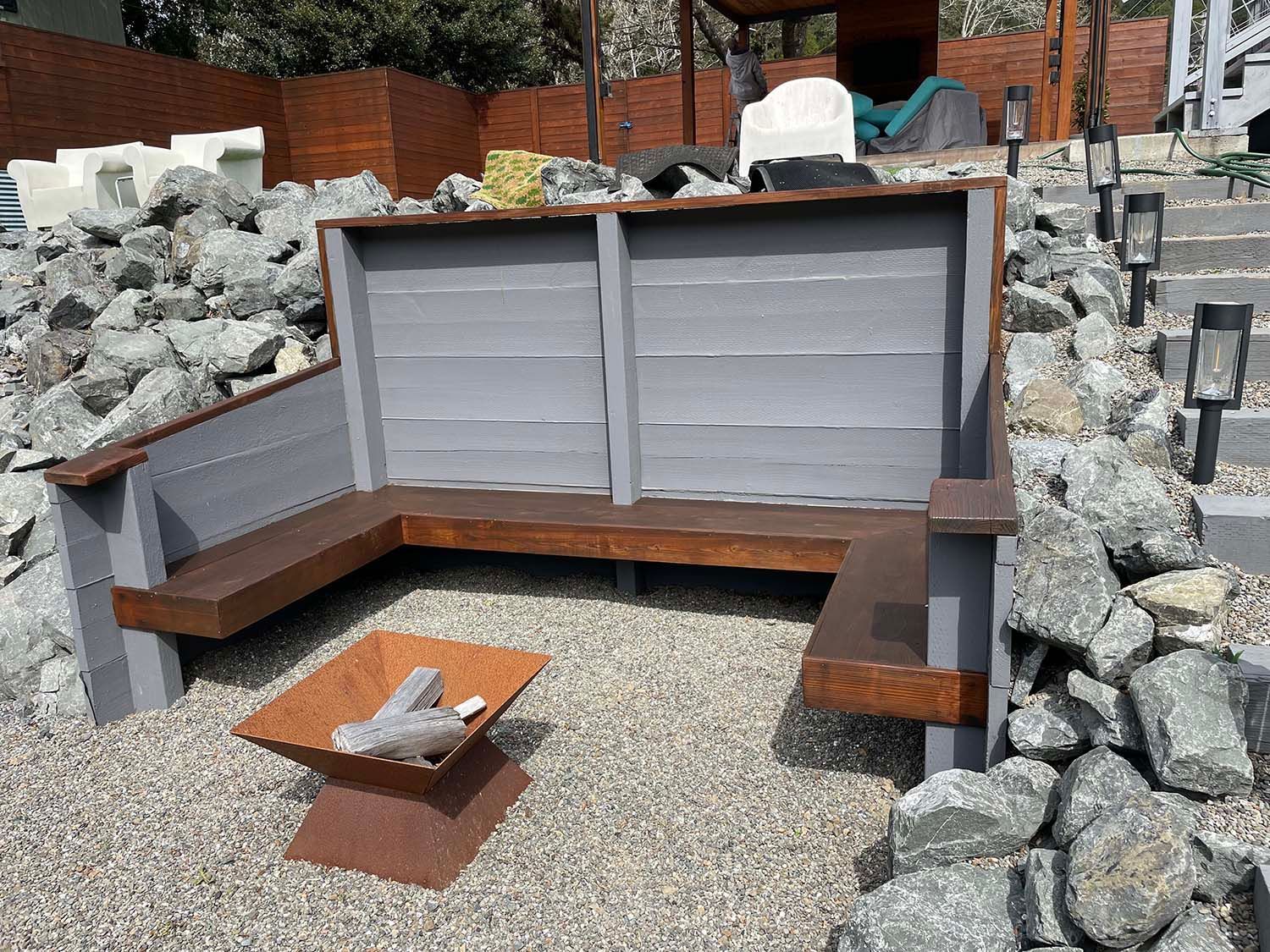L-shaped wooden bench around a gravel fire pit in a landscaped outdoor seating area