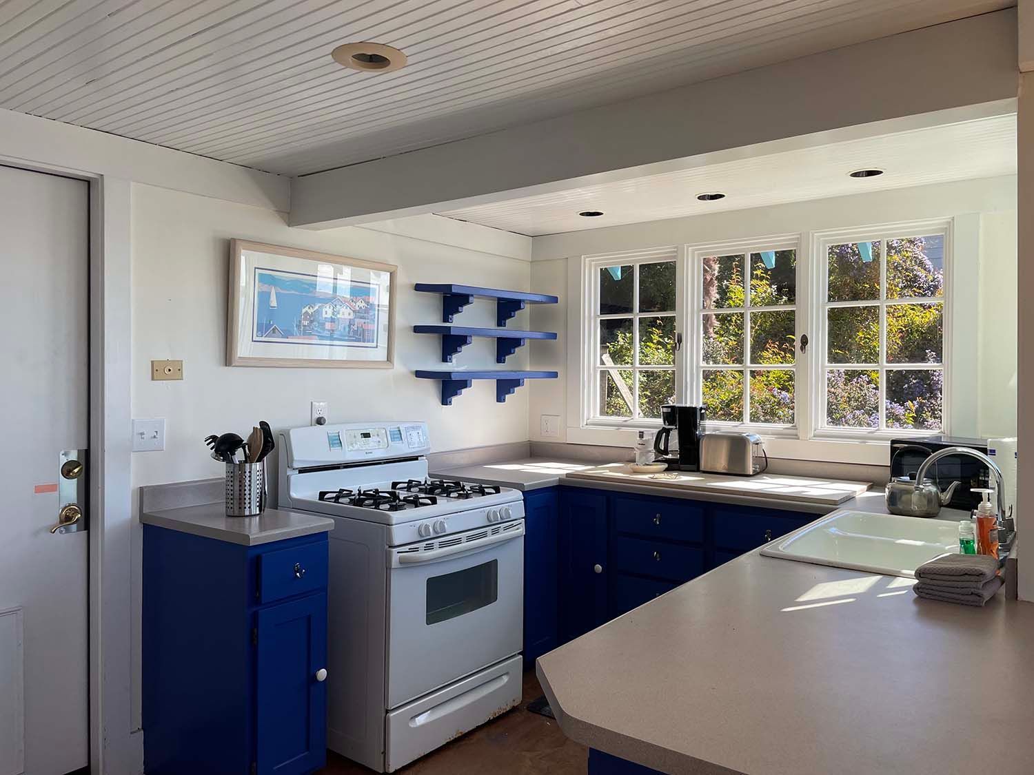 Bright kitchen with blue cabinets, white stove, large windows, and a beige countertop