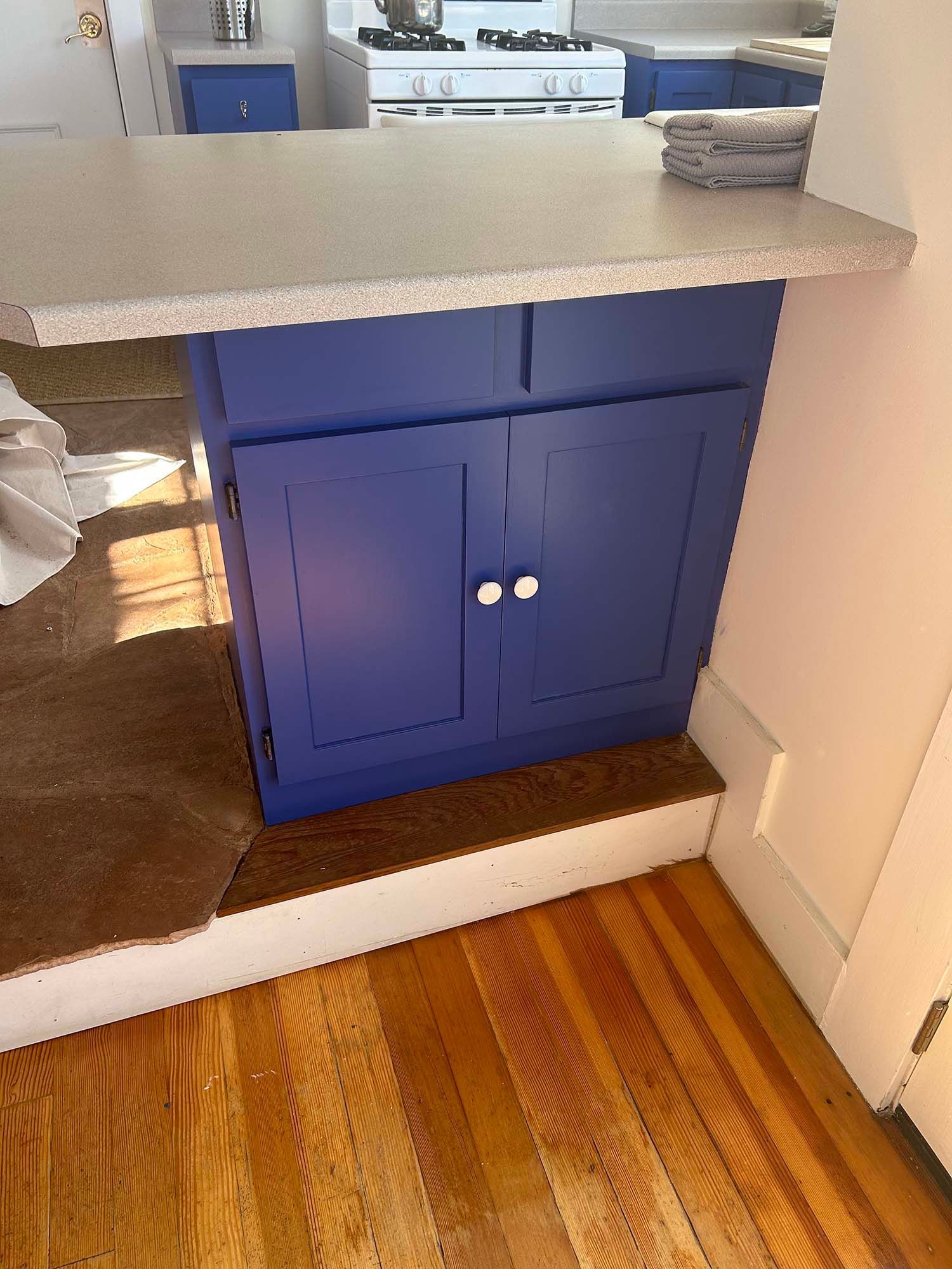 Blue kitchen cabinet under a speckled countertop, next to a hardwood floor and white baseboard.