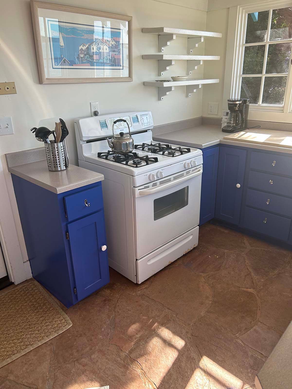 Small kitchen with white stove, blue cabinets, shelves, sink, and sunlight on the floor.