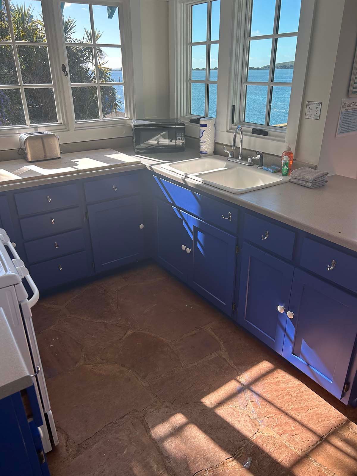 Bright blue kitchen with white countertops, sunlit corner windows, and terracotta tile floor