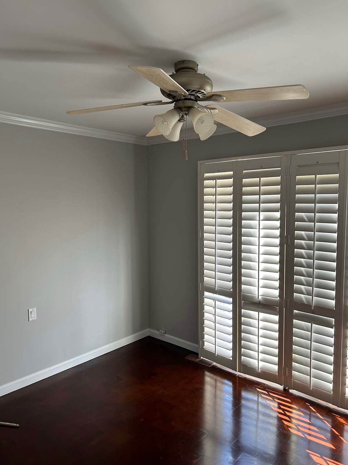 Empty room with gray walls, dark hardwood floor, ceiling fan, and white plantation shutters.