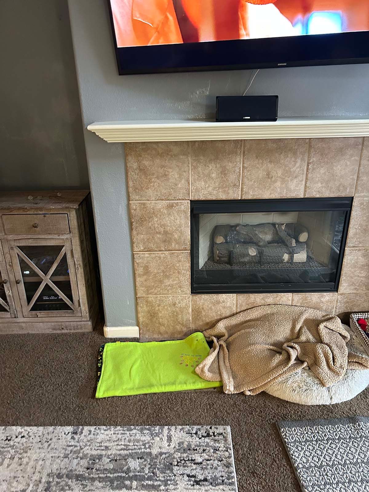 Living room fireplace with TV above, green blanket and beige pet bed on the floor